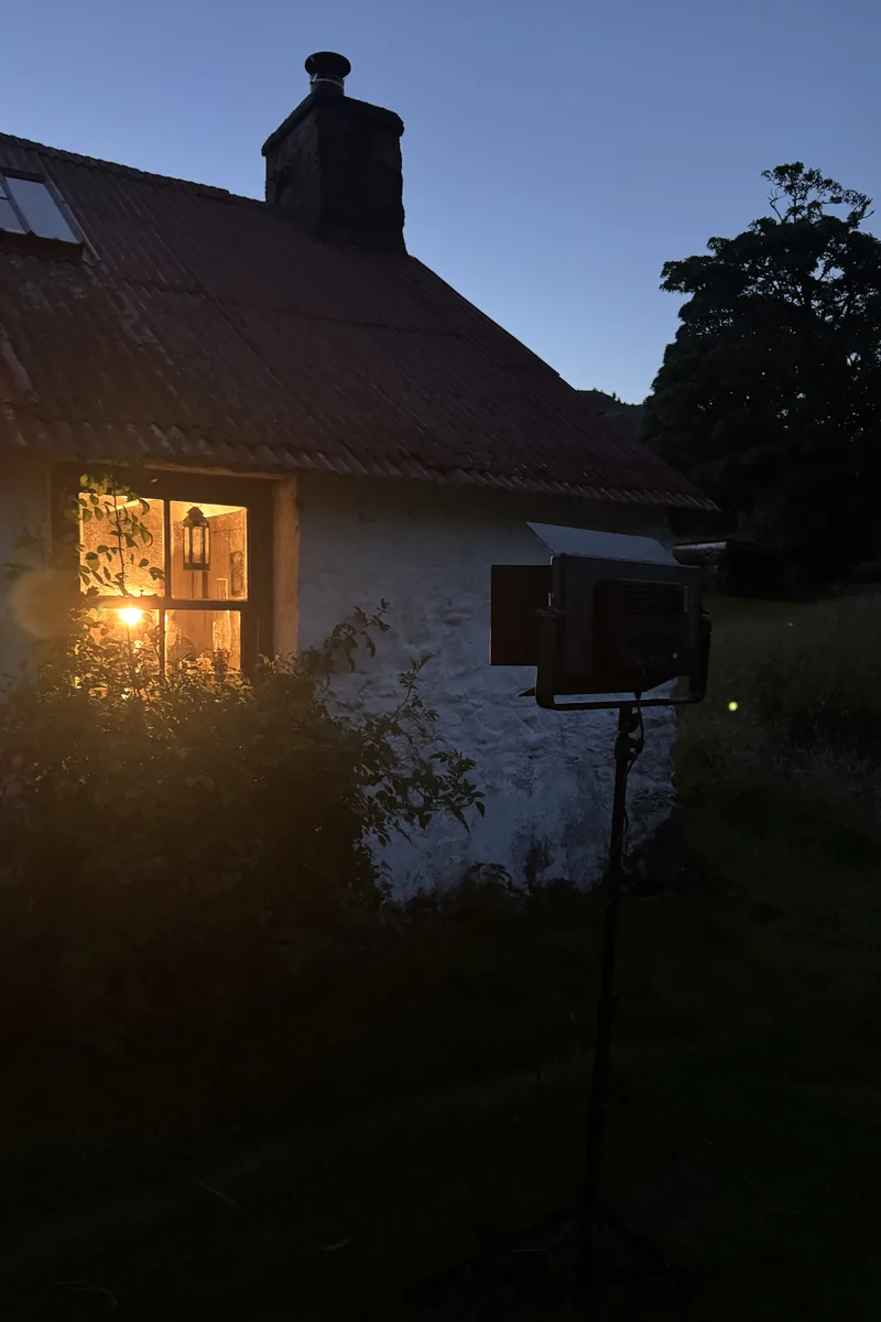 Nächtlicher Drehaufbau: Filmleuchte auf einem Stativ vor einem Cottage, warmes Licht im Fenster