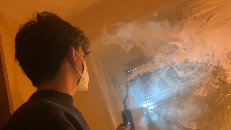 Luis mit Maske hält ein Licht in Rauch vor einem Fenster