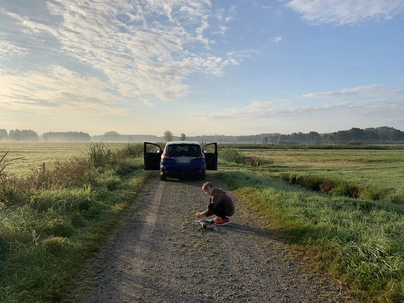 Luis kniet auf einem Feldweg neben seiner Drohne, dahinter das Auto