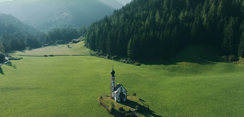 Luftaufnahme: die Kirche St. Johann in Ranui auf einer grünen Wiese vor Bergwald